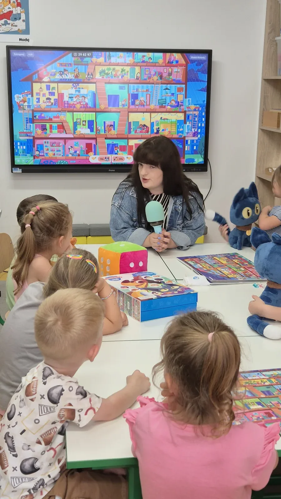 Nauczycielka przy stole z mikrofonem prowadzi zajęcia z grupą dzieci